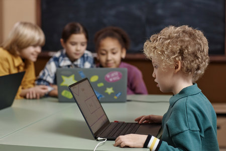 Side view portrait of young curly haired boy using laptop in school during IT class for little kids...