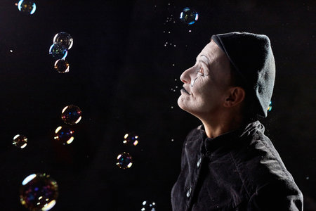 Minimal profile view portrait of mime artist looking at soap bubbles against black background, copy...