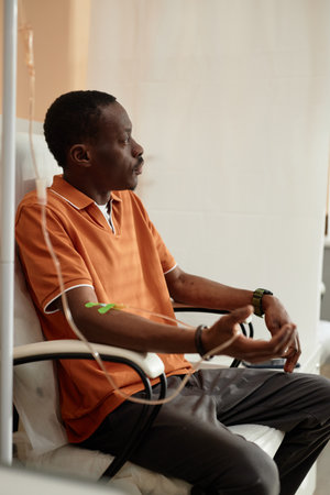 Vertical side view portrait of pensive African American man getting IV drip treatment sitting in...