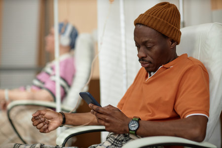 Side view of adult Black man using mobile phone during chemotherapy treatment in clinic copy spaceの写真素材