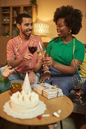 Vertical portrait of Black young woman clinking wine glasses with friend during cozy Birthday partyの写真素材