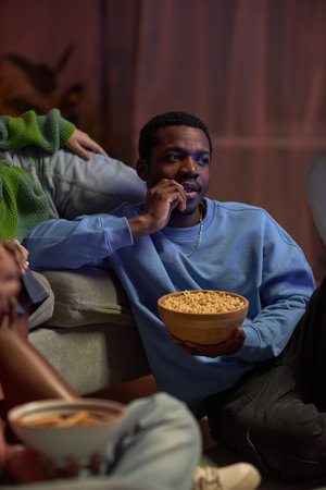 Vertical side view portrait of adult African American man watching movie with friends sitting on floor and eating popcornの写真素材