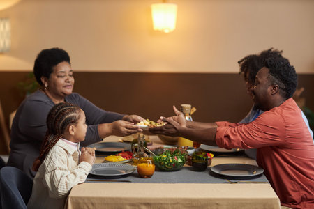Side view portrait of three generation African American family enjoying dinner together in cozy home and sharing food over tableの写真素材