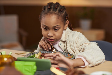 Front view of little African American girl using smartphone and watching videos while eating at dinner tableの写真素材
