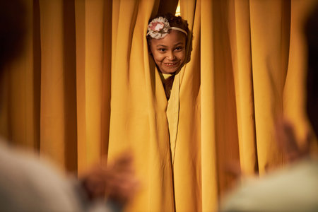 Portrait of little Black girl with head peeking through yellow curtains on stage in theater copy spaceの写真素材