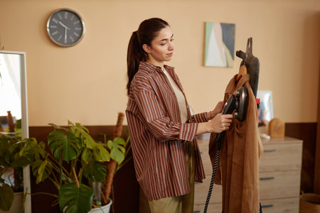 Side view portrait of young woman using garment steamer on velvet jacket in cozy home copy space