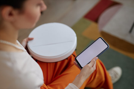 High angle closeup of young woman setting up robot vacuum cleaner and holding smartphone with white...