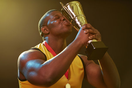 Waist up portrait of muscular Black man kissing trophy cup and celebrating victory in sports competitionの写真素材