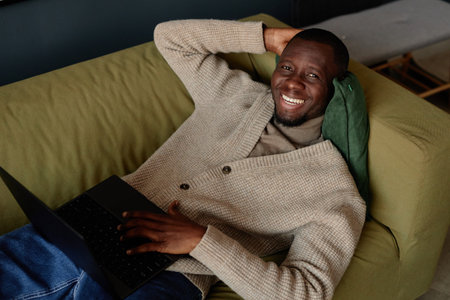 High angle portrait of smiling African American man lying on couch with laptop and looking at camera while relaxing at workの写真素材