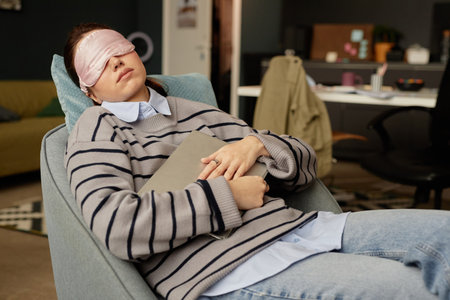 Portrait of young woman sleeping in armchair with sleep mask on while taking break from work