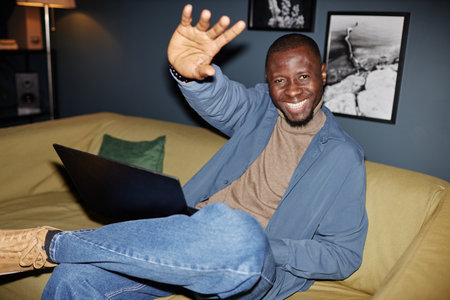 Portrait of carefree Black businessman smiling at camera sitting on couch with laptop, shot with...