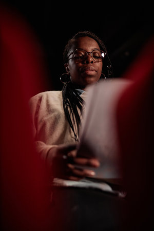 Vertical low angle portrait of African American woman reading script sitting in audience at theater...