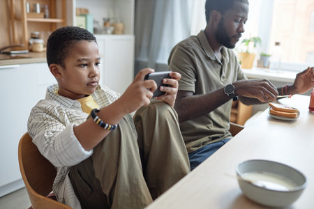 Portrait of teenage African American boy using smartphone at kitchen table during breakfast with family copy spaceの写真素材