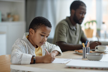 Side view portrait of African American boy doing homework at kitchen table with father working in...