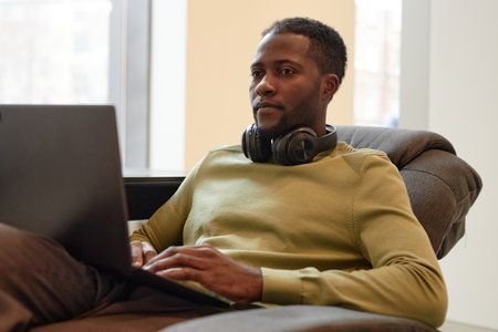 Clean portrait of young African American man using laptop and working while relaxing in office loungeの写真素材
