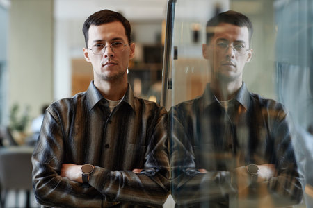 Waist up portrait of confident Asian man wearing glasses leaning on glass wall in office with...