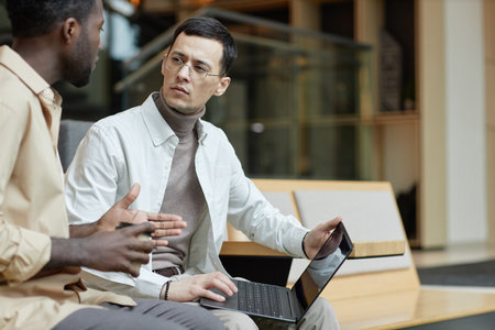 Side view portrait of Asian man wearing glasses listening to colleague discussing project and using...