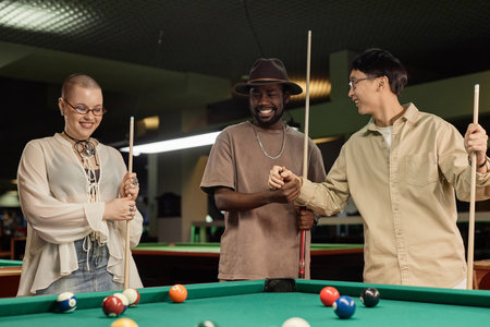 Waist up portrait of diverse group of friends enjoying game of pool together at billiards table and...