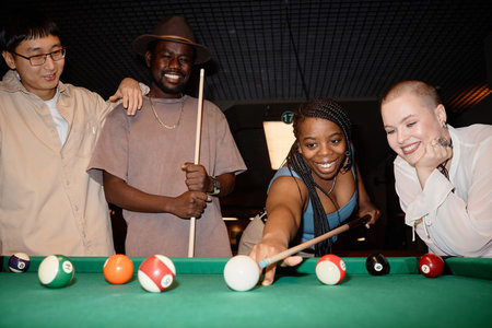Multiethnic group of smiling young people playing pool together with Black woman hitting ball with...