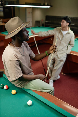 Vertical portrait of Black adult man wearing hat posing on pool table in barの写真素材
