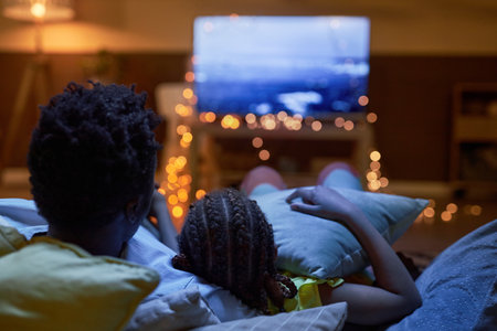 Back view of Black family watching Tv together in dark room sitting on sofa at homeの写真素材
