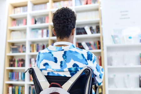 Back view of student using wheelchair with backpack looking at shelves in school library copy space