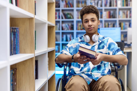 Front view portrait of teenage boy with disability using wheelchair in school library and holding books looking at camera, copy spaceの写真素材
