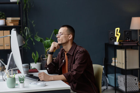 Side view portrait of young Asian man wearing glasses and thinking while looking at computer screen...