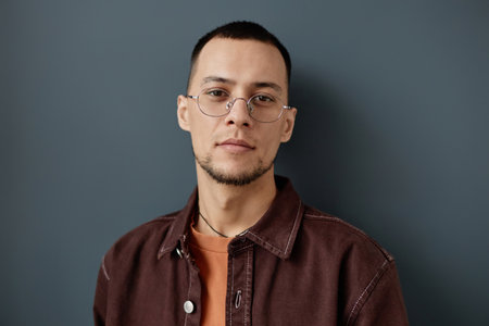 Minimal portrait of adult Asian man wearing glasses standing against grey wall dressed in casual...