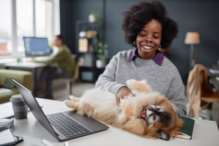 Portrait of smiling African American woman playing with cute dog while working at desk in pet...