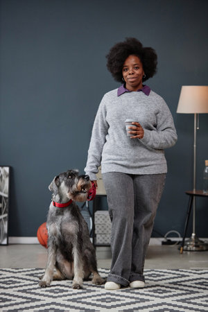 Vertical full length portrait of smiling African American woman with schnauzer dog looking at camera...
