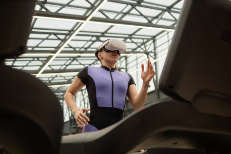 Graphic low angle portrait of sportive woman wearing VR headset and running on treadmill in futuristic gym against glass ceilingの写真素材