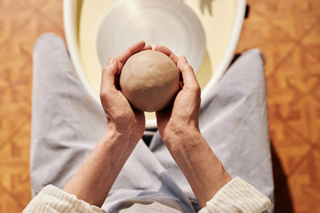 Minimal top view closeup of woman gently holding clay ball in hands over pottery wheel in sunlit art studio copy spaceの写真素材