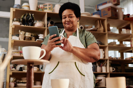 Low angle portrait of African American senior woman using smartphone in pottery studio and posting...