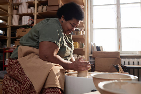 Side view portrait of smiling senior African American woman creating handmade ceramics on pottery wheel and enjoying cozy hobby in art studio copy spaceの写真素材