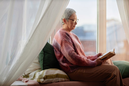 Side view portrait of senior woman reading book while relaxing by window alcove and enjoying retirement copy spaceの写真素材