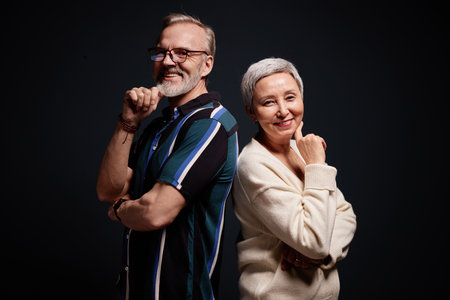 Waist up portrait of two smiling senior people standing back to back and looking at camera in studio
