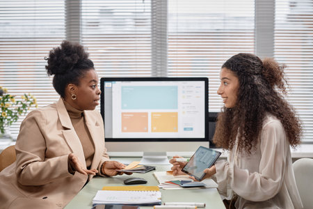 Side view of two businesswomen discussing new mobile app interface design while holding color swatches. Diverse team talk shop against window in office, copy spaceの写真素材