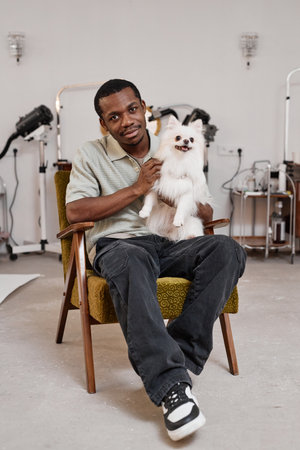 Vertical full length portrait of young Black man holding cute Spitz dog in lap while sitting on chair in grooming studioの写真素材