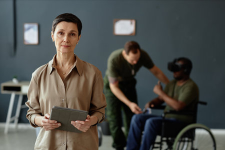 Waist up portrait of mature Caucasian woman as mental health expert looking at camera holding tablet...