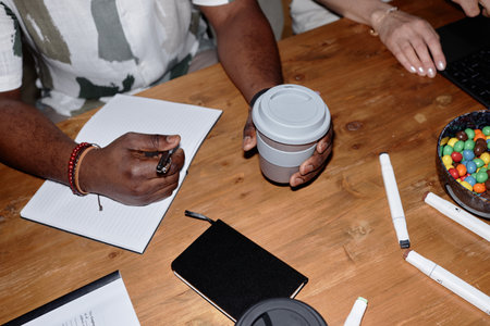 Top view closeup of unrecognizable Black man taking notes and holding coffee cup sitting at wooden...