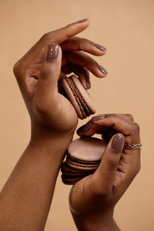 Vertical close up of elegant female hands with dark brown manicure holding cappuccino macaron in matching color on beige background. Nail design ideas for chocolate loversの写真素材