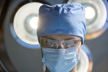 Low angle close up of focused male surgeon wearing mask and protective glasses looking down under lamp in operating room copy spaceの写真素材