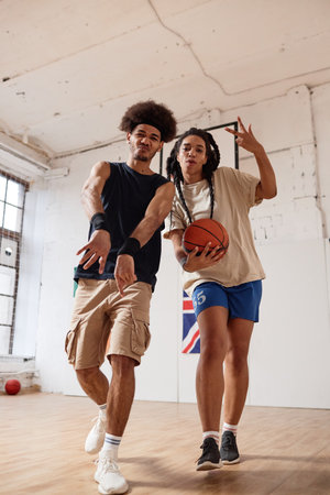 Vertical full length shot of two Black teenagers boy and girl playing basketball together and...