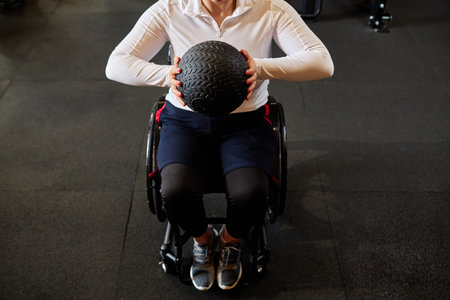 Top view closeup of unrecognizable woman using wheelchair in gym and holding medball during strength...