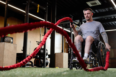 Motivational full length portrait of man with disability using wheelchair while training with battle rope in gym, copy spaceの写真素材
