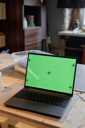 Displaying laptop with green screen placed on wooden desk in modern workspace featuring shelves with office supplies and window in backgroundの写真素材