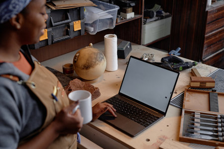 Woman working in workshop holding mug while looking at laptop for design work surrounded by various tools and materialsの写真素材