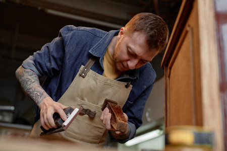 Woodworker concentrating on sanding wooden furniture piece, wearing apron and surrounded by workshop tools and equipment. Tattoed arms visible while engaged in meticulous craftsmanshipの写真素材