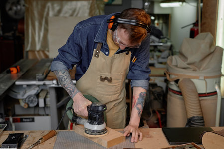 Person seen concentrating on sanding wood piece with sander machine in workshop filled with various tools and gadgets surrounding. Wearing protective gear and focused on taskの写真素材